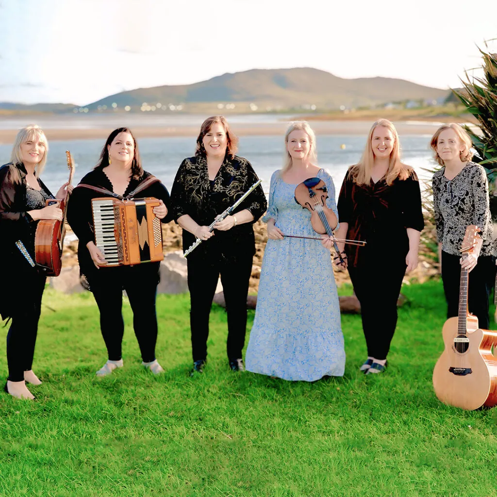 Six women stand on a green lawn, holding various musical instruments, with a scenic water and hill view in the background.