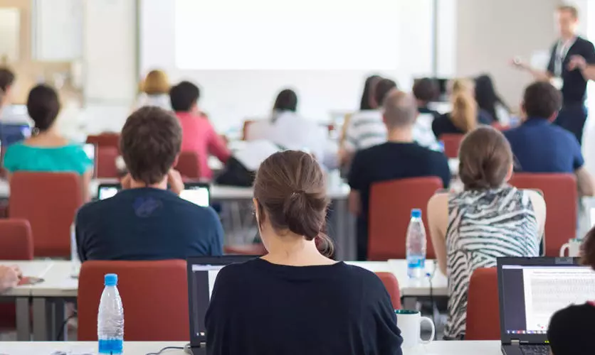 Photo of a classroom from the back where you can see the backs of students' heads and the professor at the front of the room.